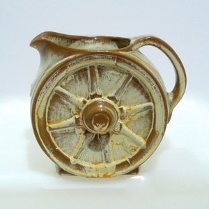 May include: A ceramic pitcher with a wagon wheel design. The pitcher is primarily beige with brown accents, including the handle, spout, and wheel details. The wheel is prominently featured on the side.