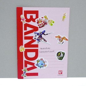 May include: A pink Bandai catalog from 2008 with a red spine. The cover is decorated with various stickers, including a robot, a green watch, and several small cartoon figures. The Bandai logo is prominently displayed.