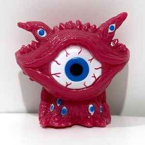 May include: A magenta-colored monster figurine with a large central eye and multiple smaller blue eyes. The figurine has a textured surface, sharp teeth, and pointed horns. The eye has a white iris, blue pupil, and red veins.
