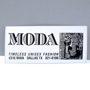 May include: A white rectangular card with the word "MODA" in large, black, block letters. Below, the text reads "TIMELESS UNISEX FASHION" and an address and phone number. A black and white illustration of an Egyptian pharaoh is on the right.