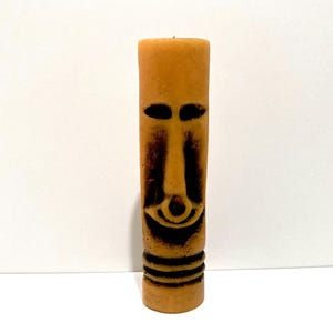 May include: A tall, cylindrical candle with a carved face design. The candle is a light brown color with dark brown accents that define the facial features. The face has oval eyes, a long nose, and a smiling mouth. The base has three horizontal lines.