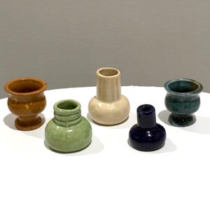 May include: A collection of five small ceramic vessels in various shapes and colors. The set includes a mustard yellow pedestal vase, a light green ribbed vase, a cream-colored bottle vase, a dark blue bottle vase, and a teal pedestal vase.