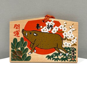 May include: A wooden ema plaque featuring a brown wild boar illustration centered over a large red circle. The design includes white plum blossoms, green pine needles, and black Japanese kanji characters. An orange cord is attached at the top.