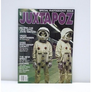May include: A copy of Juxtapoz Art & Culture Magazine's Special Photography Issue. The cover features two figures in beige space suits with skull-like visors. The magazine's title is in large purple letters. The cover also includes text about John Waters and Viggo Mortensen.