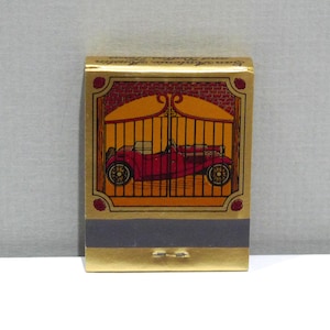 May include: A gold matchbox with a red vintage car illustration behind a gate. The matchbox has a dark grey strip with two match heads. The top of the matchbox has text in gold.