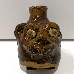 May include: A handmade, brown ceramic jug with a whimsical face design. The jug features large, round eyes, a wide nose, and a smiling mouth. The jug is approximately 4 inches tall and has a glossy finish.