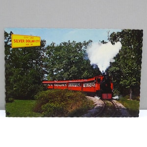 May include: Vintage postcard featuring a red train with a white smoke plume, traveling through a green landscape. The postcard has a scalloped edge and a yellow sign that reads "Silver Dollar City".