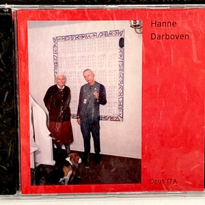 May include: A CD case with a red cover featuring the name "Hanne Darboven" and "Opus 17A." The cover art shows two people and a dog in a room with a patterned wall hanging. The CD is in a clear plastic case.
