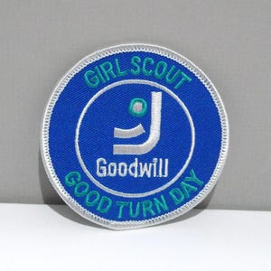 May include: A circular patch with a blue background and white stitching. The patch features the words "Girl Scout Good Turn Day" in white and green, surrounding a white and green Goodwill logo.