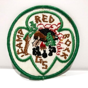 May include: Embroidered patch with a green border and the words "Camp Red Rock GS" in brown. The patch features a cabin, trees, and a campfire in the center, with a white background.