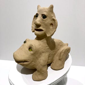 May include: A tan ceramic sculpture of a creature with a human-like face atop an animal-like body. The sculpture has green glass eyes and is sitting on a white surface. The artwork is a unique piece of home decor.