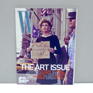 May include: A magazine cover featuring a woman holding a sign that reads "IT MUST BE SOMEBODY'S FAULT." The cover has the "W" logo and the text "THE ART ISSUE." The background shows a city street with a yellow taxi and people.