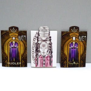 May include: Three perfume bottles and boxes. The boxes are brown with gold accents and the word "ALIEN" in white. The perfume bottle in the center has a silver face design and a chain. The perfume inside is pink.