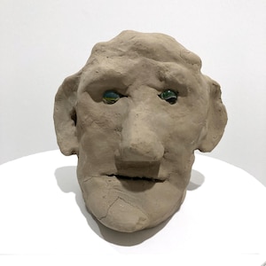 May include: A handmade, light brown clay sculpture of a human face. The face has large ears, a prominent nose, and green glass eyes. The sculpture is on a white surface.