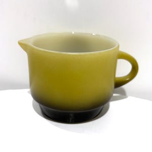 May include: A vintage, olive-green creamer with a handle and a spout. The creamer has a rounded body that sits on a dark base. The interior is a lighter shade of green.