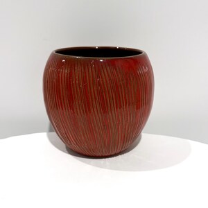 May include: A round ceramic planter with a glossy red glaze and a textured surface featuring vertical, thin, brown etched lines. The vessel has a wide, open top and a smooth, dark brown interior rim, showcasing a consistent, rounded shape.