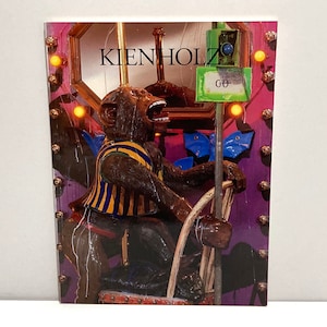 May include: A print featuring a monkey sculpture with an open mouth, wearing a striped vest and holding a steering wheel. The word "KIENHOLZ" is visible at the top. The background is a mix of purple, gold, and blue.