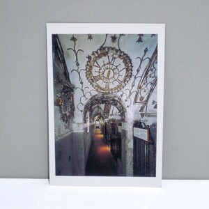 May include: A postcard featuring a long, arched hallway with a clock on the ceiling. The clock has Roman numerals and is surrounded by ornate floral designs. The hallway is lit with warm light.