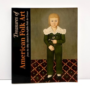 May include: A hardcover book titled Treasures of American Folk Art from the Abby Aldrich Rockefeller Folk Art Center. The cover features a painting of a child in a dark green suit with a white ruffled collar, holding a small bird in one hand.