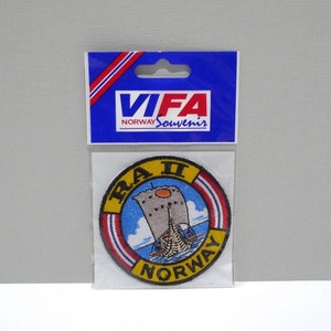 May include: A round embroidered patch featuring a ship with a sail, the text "RA II NORWAY" in yellow, and a blue sky background. The patch is packaged in a clear plastic sleeve with a blue header that reads "VIFA NORWAY Souvenirs".