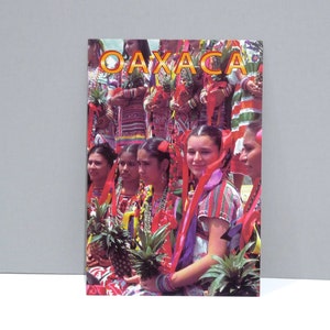 May include: A postcard featuring a group of women in traditional Oaxacan attire. The women are holding pineapples and wearing colorful dresses with red ribbons in their hair. The word "OAXACA" is printed in orange letters at the top.