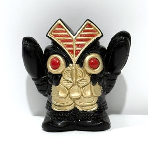 May include: A black figurine with a gold and red design. The figurine has large, red eyes and a gold chest. The head has a red and gold design. The figurine has two black arms extending from the sides.