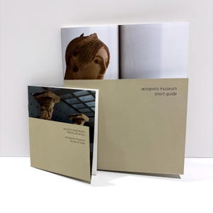 May include: A beige Acropolis Museum short guide booklet and a smaller museum plan pamphlet. The guide features a photograph of an ancient stone sculpture head, while the plan shows architectural columns with the text Acropolis Museum.