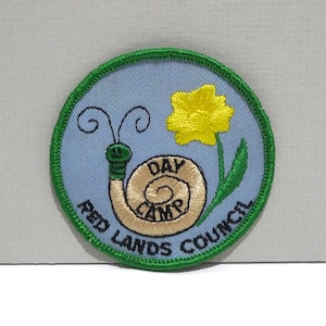 May include: Embroidered patch with a light blue background, featuring a yellow flower and a snail with the words "DAY CAMP" in its shell. The patch is bordered with green stitching and includes the text "RED LANDS COUNCIL".