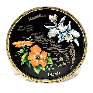 May include: Round black tray with a gold rim featuring a map of the Hawaiian Islands. The tray has orange hibiscus flowers and white orchids. The word "Hawaiian" is at the top, and "Islands" at the bottom.