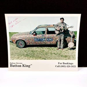 May include: A vintage photograph featuring a man in a button-covered suit, holding a guitar, standing next to a car covered in buttons. The car has the text "BUTTON KING" on the side. A banjo rests on the ground. The photo includes the text "Dalton Stevens Button King™".