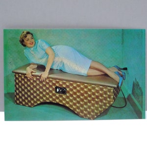 May include: Vintage postcard featuring a woman in a light blue dress reclining on a gold-colored, textured bench. The bench has a control panel and is set against a teal background. The woman is wearing blue heels.