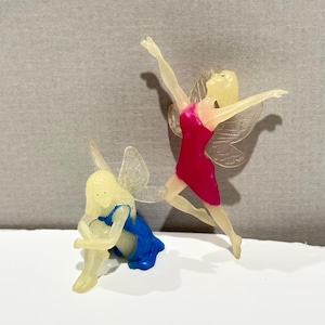 May include: Two small fairy figurines made of translucent off-white material. One fairy is in a kneeling position wearing a blue dress, while the other is in a leaping pose with arms extended upward, wearing a bright pink dress and translucent wings.