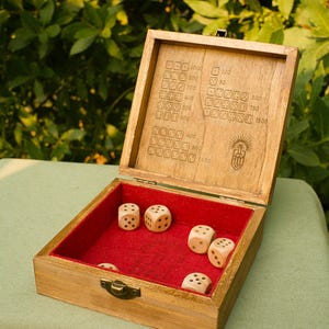 May include: A wooden dice game box with a hinged lid featuring an engraved scoring chart and a logo. The interior is lined with red felt and contains six light-colored wooden dice. The box has a brass-colored metal latch on the front edge.