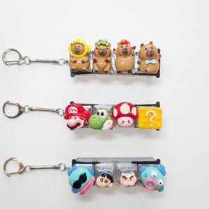 May include: Three metal keychains featuring rows of four small, colorful, three-dimensional character figures attached to clear plastic blocks. The top row shows brown capybara figures, the middle row features video game characters, and the bottom row displays various cartoon faces.
