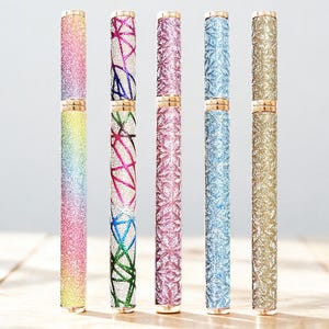 May include: Six glitter eyeliner pens in various colors, including rainbow, pink, blue, and gold. Each pen has a gold-colored cap and base. The pens are arranged vertically on a light-colored wooden surface.