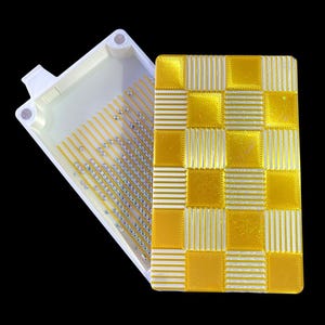 May include: A white and yellow plastic container with a lid. The lid has a checkered pattern of yellow squares and white stripes. The container has a series of small, silver-colored beads. The container is open, revealing the interior.
