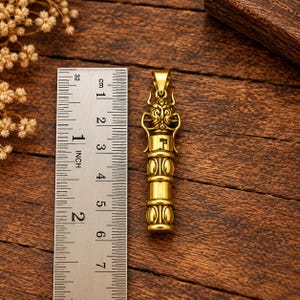 Puede incluir: Un colgante de metal dorado con forma de criatura estilizada con cuernos, junto a una regla metálica que muestra una longitud de aproximadamente 5,7 cm. El colgante presenta un cuerpo cilíndrico con bandas decorativas y una anilla superior.