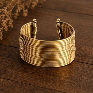 Puede incluir: Un brazalete ancho tipo puño construido con múltiples anillos de alambre apilados en tono dorado. El diseño presenta una serie continua de bandas metálicas horizontales que forman una estructura cilíndrica abierta, con detalles de espirales verticales.