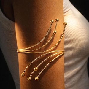 Puede incluir: Un brazalete dorado para el brazo que presenta una serie de hilos metálicos delgados y curvos que se abren en abanico desde un punto central. Cada hilo termina en una pequeña cuenta esférica pulida, creando un diseño lineal y radiante.
