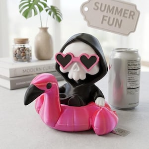 May include: A figurine of a white skull wearing a black hooded cloak and pink heart-shaped sunglasses, sitting inside a bright pink inflatable flamingo pool float. A tag above the figure displays the text SUMMER FUN in a simple font.