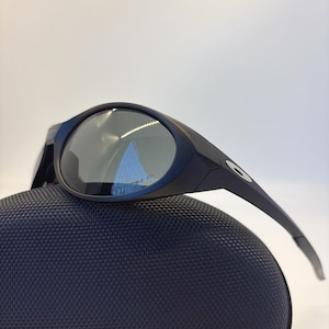 May include: Black sunglasses with a curved frame and dark lenses rest on a textured, navy blue case. The sunglasses have a sleek, modern design, with a small logo on the temple.