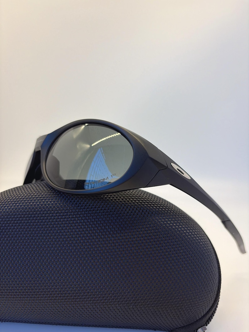 May include: Black sunglasses with a curved frame and dark lenses rest on a textured, navy blue case. The sunglasses have a sleek, modern design, with a small logo on the temple.