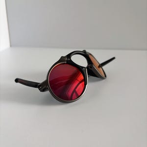 May include: Round sunglasses with a dark frame and red lenses. The sunglasses have a classic design with a metal frame and a bridge. The lenses are a vibrant red color, and the frame is a dark color.