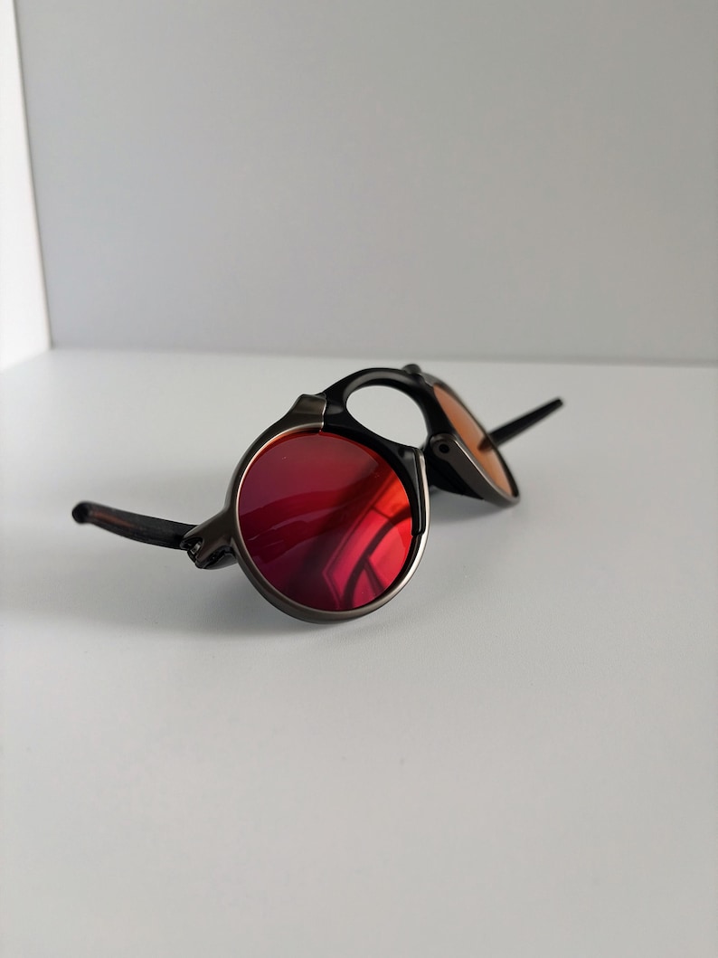 May include: Round sunglasses with a dark frame and red lenses. The sunglasses have a classic design with a metal frame and a bridge. The lenses are a vibrant red color, and the frame is a dark color.
