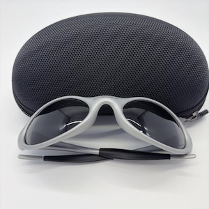 Puede incluir: Gafas de sol con montura plateada y lentes oscuras, frente a un estuche negro y texturizado para gafas de sol. Las gafas de sol tienen un dise&ntilde;o elegante y moderno. El estuche es de forma ovalada con cierre de cremallera.