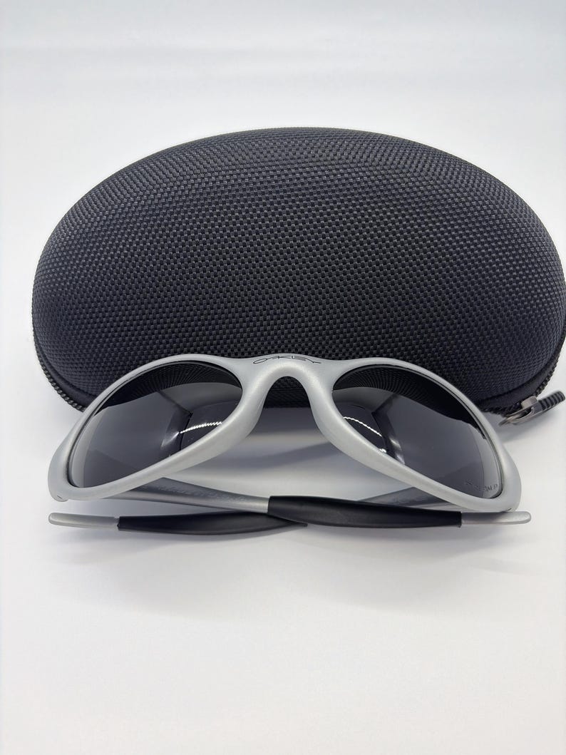 Puede incluir: Gafas de sol con montura plateada y lentes oscuras, frente a un estuche negro y texturizado para gafas de sol. Las gafas de sol tienen un dise&ntilde;o elegante y moderno. El estuche es de forma ovalada con cierre de cremallera.
