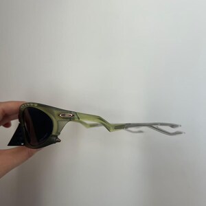 May include: Green-tinted sunglasses with dark lenses. The frames have a unique, angular design and a matte finish. The sunglasses are held by a hand, with a black tag attached to the frame.