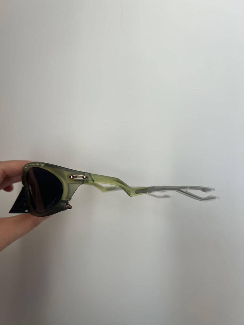 May include: Green-tinted sunglasses with dark lenses. The frames have a unique, angular design and a matte finish. The sunglasses are held by a hand, with a black tag attached to the frame.