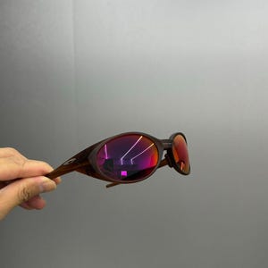 Puede incluir: Gafas de sol marrones con lentes espejadas rojas. Las gafas tienen una montura y patillas curvas. Las lentes reflejan un vibrante tono rosa y morado. Las gafas son sostenidas por una mano sobre un fondo gris.