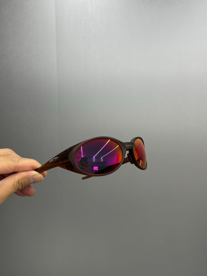 Puede incluir: Gafas de sol marrones con lentes espejadas rojas. Las gafas tienen una montura y patillas curvas. Las lentes reflejan un vibrante tono rosa y morado. Las gafas son sostenidas por una mano sobre un fondo gris.